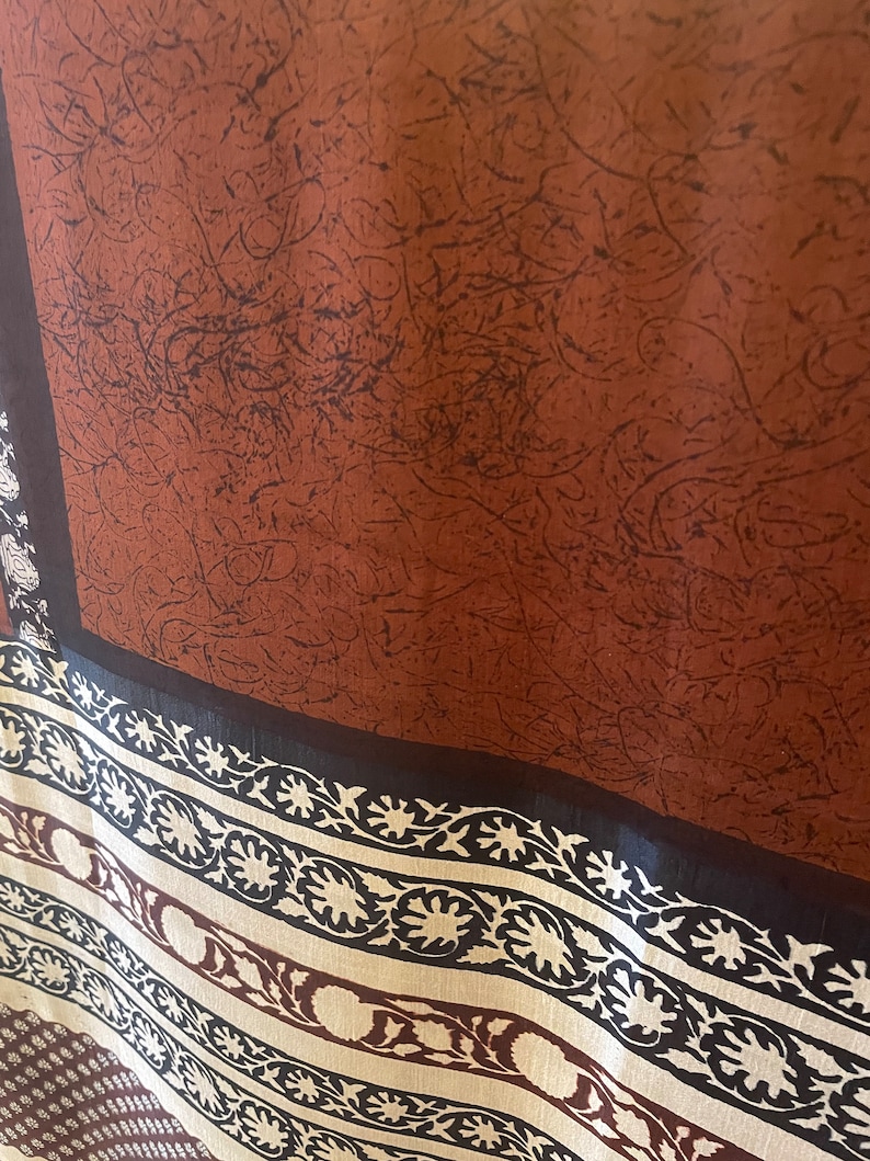 May include: A brown textile with a dark pattern and a decorative border. The border features a repeating floral design in cream and brown against a black background. The main section has a textured appearance.
