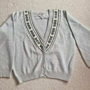 May include: A light gray cardigan sweater with a V-neck and long sleeves. The neckline is decorated with a cream and navy blue embroidered pattern. The sweater has a button-up front and ribbed cuffs and hem.