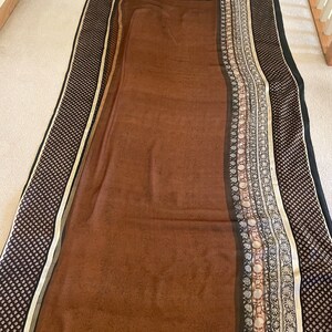 May include: A brown textile with a patterned border and a central field. The border features a repeating dot pattern in black and white, with a stripe of gold and cream. The central field is a solid brown color with a subtle pattern.