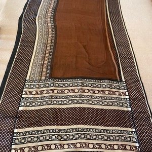 May include: A long, rectangular scarf with a brown center and patterned borders. The borders feature black, white, and brown geometric and floral designs. The scarf has a black border on the edges.