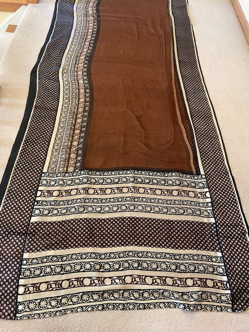 May include: A long, rectangular scarf with a brown center and patterned borders. The borders feature black, white, and brown geometric and floral designs. The scarf has a black border on the edges.