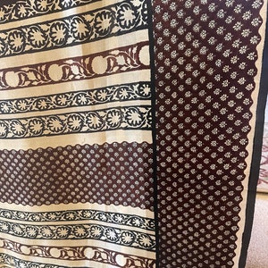 May include: A patterned textile featuring alternating stripes and a floral design. The stripes alternate between a cream color with black and brown floral patterns and a dark brown background with small white flowers. The edges are trimmed with a black border.