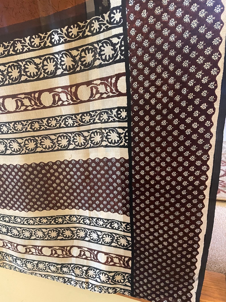 May include: A patterned textile featuring alternating stripes and a floral design. The stripes alternate between a cream color with black and brown floral patterns and a dark brown background with small white flowers. The edges are trimmed with a black border.