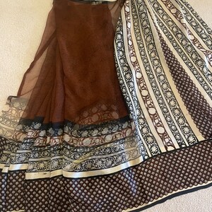 May include: A brown and white patterned textile with a sheer overlay. The textile features a variety of patterns, including small dots, floral designs, and stripes. The edges are trimmed with black and white borders. The fabric is draped on a light-colored surface.
