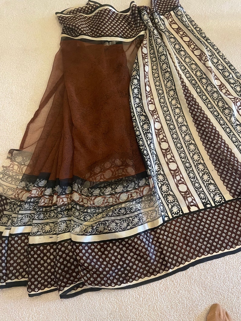 May include: A brown and white patterned textile with a sheer overlay. The textile features a variety of patterns, including small dots, floral designs, and stripes. The edges are trimmed with black and white borders. The fabric is draped on a light-colored surface.