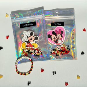 May include: Two clear plastic bags with Mickey Mouse and Minnie Mouse character stickers. Each bag contains colourful beads and a bracelet with the name "Georgia" and "Olivia" spelled out in white beads.