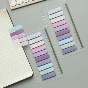 May include: Three sets of colourful sticky notes with rounded edges. Each set has a ruler on the side and is arranged in a rainbow colour scheme of light purple, pink, lavender, blue, and light blue.