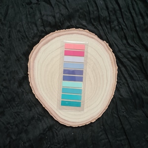 May include: A set of colourful sticky notes in a variety of shades of blue, pink, and purple. The notes are arranged in a vertical stack on a wooden surface.