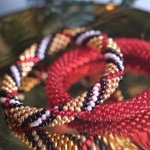 Peut inclure: Deux bracelets de perles. L'un est rouge, l'autre est un mélange de perles rouges, dorées, noires et blanches. Les bracelets sont présentés sur une surface dorée réfléchissante. Les bracelets sont un type de bijou.