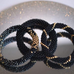 Peut inclure: Trois bracelets circulaires fabriqués à partir de petites perles noires mates. Chaque bracelet présente un motif distinct de perles dorées métalliques tissées dans le perlage noir, créant un design en spirale ou segmenté sur le bijou.