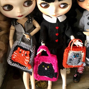 May include: Three dolls with black hair, blonde hair, and grey hair are holding crocheted cat-shaped handbags. The handbags are in pink, orange, and grey.