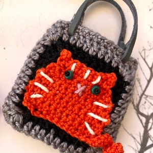 May include: A small crocheted bag with a grey and black pattern. The bag has a leather handle and features a crocheted orange cat with white stripes and green eyes.