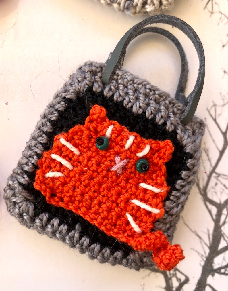 May include: A small crocheted bag with a grey and black pattern. The bag has a leather handle and features a crocheted orange cat with white stripes and green eyes.