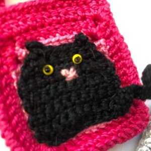 May include: A pink crocheted bag with a black crocheted cat face and tail. The cat has yellow eyes.