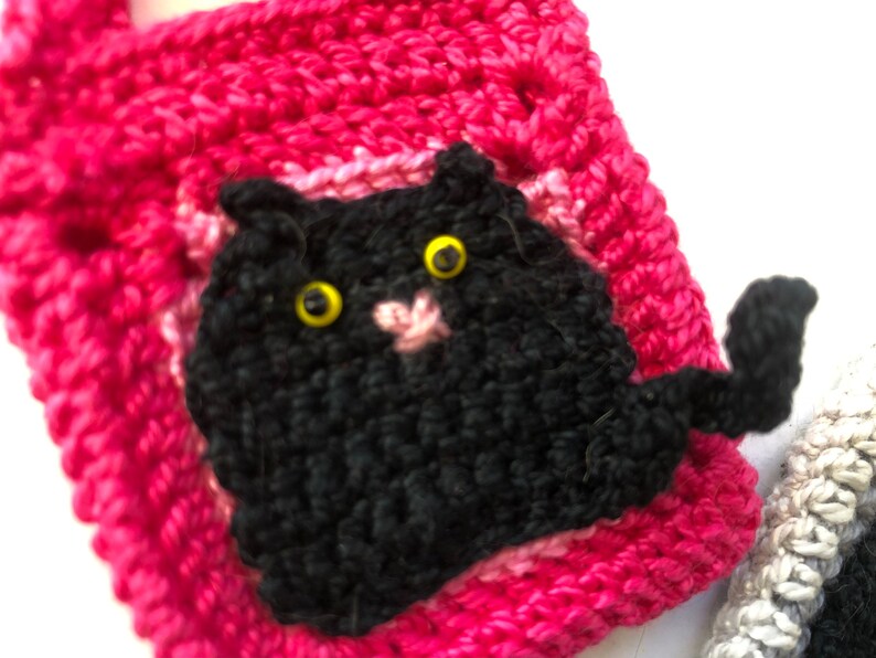 May include: A pink crocheted bag with a black crocheted cat face and tail. The cat has yellow eyes.