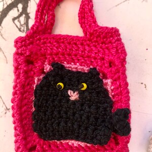 May include: A pink crocheted bag with a black crocheted cat face on the front. The cat has yellow eyes and a pink nose.