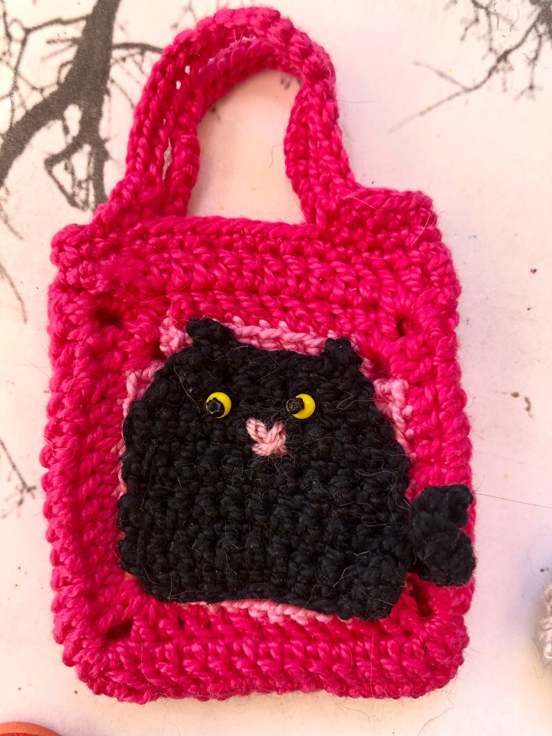 May include: A pink crocheted bag with a black crocheted cat face on the front. The cat has yellow eyes and a pink nose.