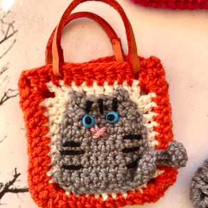 May include: A small orange crocheted bag with a grey crocheted cat face and blue eyes. The bag has brown leather straps.