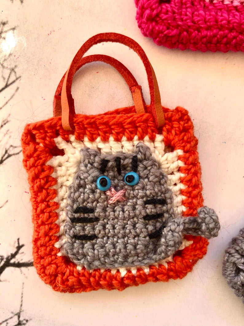 May include: A small orange crocheted bag with a grey crocheted cat face and blue eyes. The bag has brown leather straps.