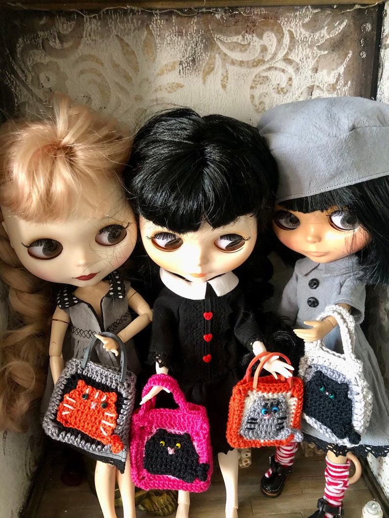 May include: Three dolls with black hair, blonde hair, and grey hair are holding crocheted cat-shaped handbags. The handbags are in pink, orange, and grey.