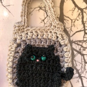 May include: A crocheted grey and white bag with a black crocheted cat with green eyes and a pink nose. The bag has a handle and is small enough to be a keychain.