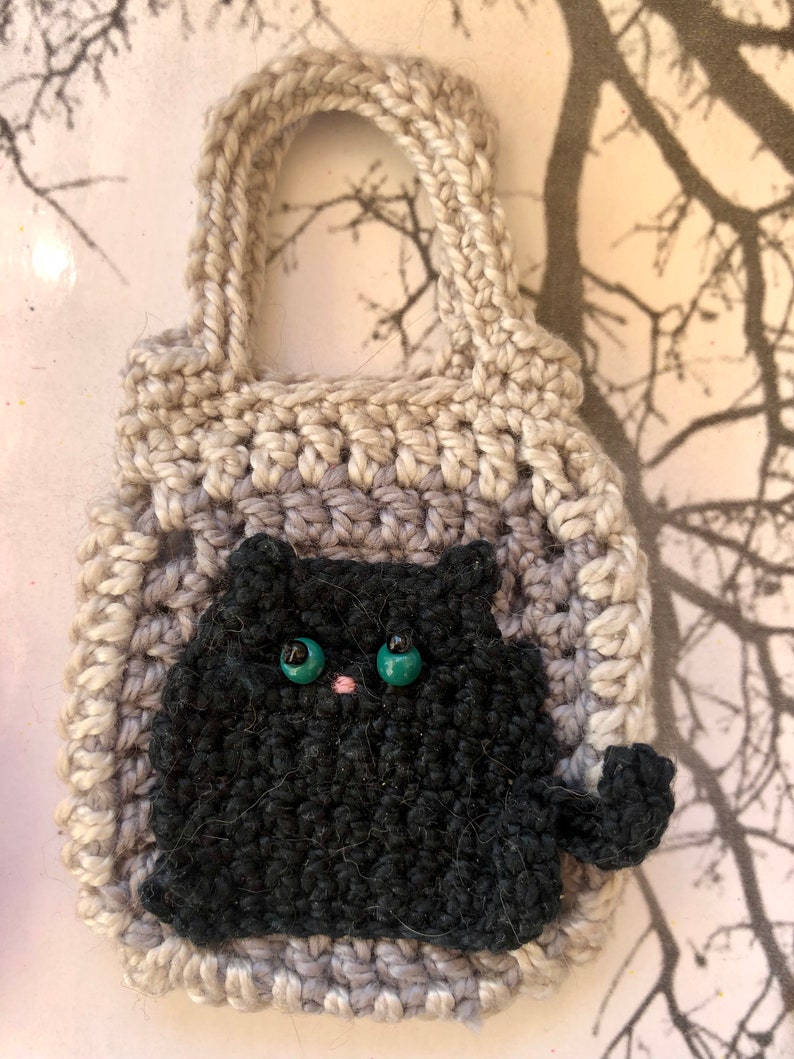 May include: A crocheted grey and white bag with a black crocheted cat with green eyes and a pink nose. The bag has a handle and is small enough to be a keychain.