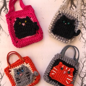 May include: Four crocheted cat-shaped bags with different coloured handles and cat faces. The bags are pink, grey, orange, and black. The cats are black, grey, orange, and black.