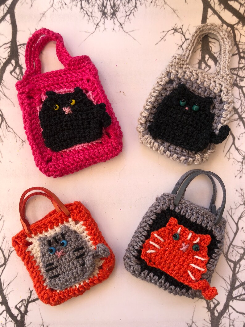 May include: Four crocheted cat-shaped bags with different coloured handles and cat faces. The bags are pink, grey, orange, and black. The cats are black, grey, orange, and black.
