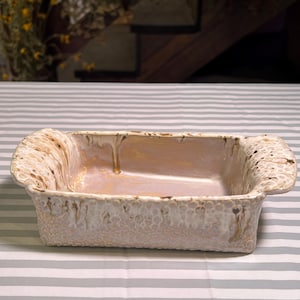 May include: Rectangular ceramic baking dish with a speckled, textured exterior in cream and tan hues. The interior has a light pinkish-beige glaze. The dish has handles on each end and sits on a gray and white striped surface.