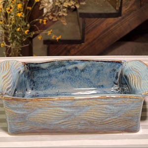 May include: A rectangular ceramic dish with a blue interior and exterior. The exterior features a wave pattern in a lighter blue and gold. The dish has rounded handles on each end.