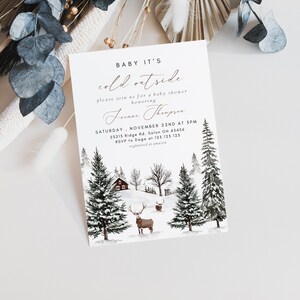 K&ouml;nnte beinhalten: Eine Babyparty-Einladung im Winter-Design mit einer Aquarell-Illustration einer verschneiten Waldszene. Der Text lautet "Baby It's Cold Outside" und enth&auml;lt Details zur Veranstaltung, wie Datum, Uhrzeit und Ort.