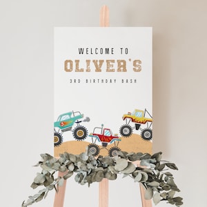 May include: Welcome to Oliver's 3rd Birthday Bash sign with a white background and brown text. The sign features three cartoon monster trucks in red, yellow, and blue colors driving on a brown dirt road.