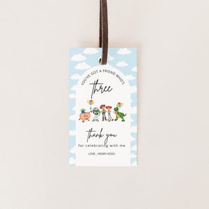 May include: A rectangular paper gift tag with a light blue sky and white cloud background. The tag features an arch design with the text "YOU'VE GOT A FRIEND WHO'S THREE" and "Thank you for celebrating with me" above "LOVE, HENRY XOXO" and cartoon characters.