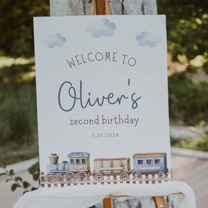 May include: A white welcome sign with blue watercolor clouds and a train illustration. The sign reads "Welcome to Oliver's second birthday 3.25.2024".