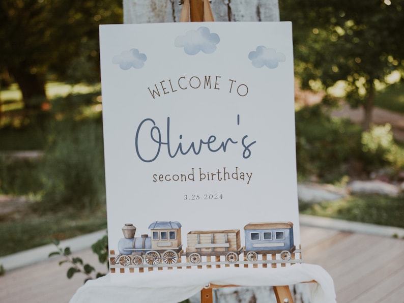 May include: A white welcome sign with blue watercolor clouds and a train illustration. The sign reads "Welcome to Oliver's second birthday 3.25.2024".
