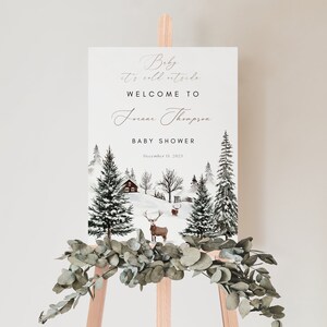 K&ouml;nnte beinhalten: Ein wei&szlig;es Schild mit einer Winterszene mit einer H&uuml;tte, B&auml;umen und Rehen. Der Text auf dem Schild lautet "Baby it's cold outside. Welcome to Jenna Thompson Baby Shower December 15, 2023."