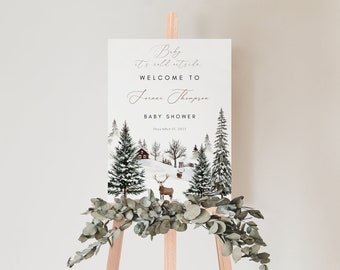 May include: A white sign with a winter scene featuring a cabin, trees, and deer. The text on the sign reads "Baby it's cold outside. Welcome to Jenna Thompson Baby Shower December 15, 2023."