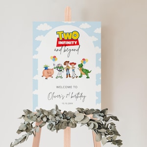 Puede incluir: Un letrero azul claro con las palabras "TWO INFINITY and beyond" en amarillo, rojo y blanco. El letrero presenta personajes de dibujos animados y el texto "Welcome to Oliver's 1st birthday". El letrero se exhibe en un caballete de madera.