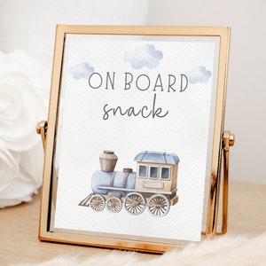 May include: A gold-framed sign with a blue and brown watercolor illustration of a train. The sign says "On Board Snack" in white text.
