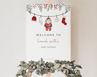 May include: Welcome to Hannah Smith's Baby Shower sign with a Christmas theme. The sign features a red and white Santa suit, a red hat, a pair of mittens, a red stocking, and a white ornament hanging from a string.