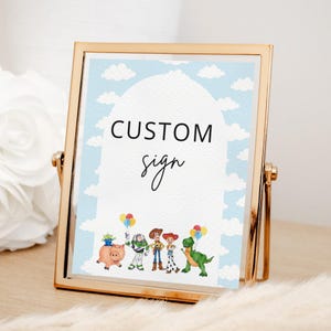 May include: A gold-toned metal frame holding a rectangular sign with a light blue sky and white cloud background. The sign features the black text CUSTOM sign and a colorful illustration of five animated toy characters holding balloons at the bottom.