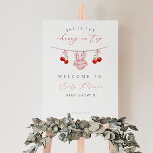 Peut inclure: Une pancarte blanche avec le texte "SHE IS THE cherry on top" en noir et rouge. Une grenouillère rose pour bébé avec des cerises rouges est suspendue à une ficelle avec des pinces à linge. Le texte "WELCOME TO Emily Peterson's BABY SHOWER" est en noir. La date "JUNE 15 20XX" est en noir.