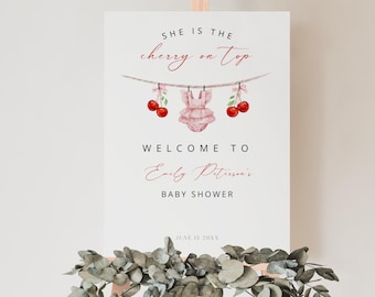 May include: A white sign with the text "SHE IS THE cherry on top" in black and red script.  A pink baby onesie with red cherries hangs from a string with clothespins.  The text "WELCOME TO Emily Peterson's BABY SHOWER" is in black script.  The date "JUNE 15 20XX" is in black text.