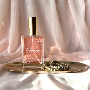 May include: A clear glass bottle with a gold spray nozzle and a pink liquid inside. The bottle is decorated with a white floral design and the text "Johnson 3/19/2024".