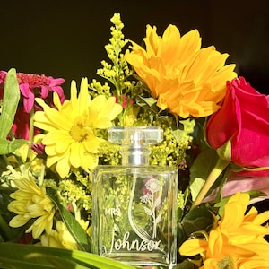 May include: A clear glass bottle of perfume with a silver cap and a floral design. The bottle is surrounded by yellow and pink flowers. The bottle is engraved with the text "MRS Johnson".