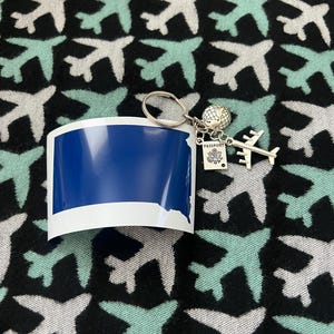 May include: A travel-themed keychain with a silver airplane charm, a globe charm, and a miniature passport tag. The keychain is paired with a blue and white sticker. The background features a pattern of airplanes in white and teal on a black background.