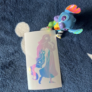 May include: A blue, purple, and orange cartoon character figurine with large eyes and colorful accents. A holographic sticker featuring a silhouette of a woman with long hair and a unicorn is also present.