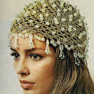 Può includere: Un copricapo all'uncinetto color oro ornato di perline trasparenti e scintillanti. Il copricapo presenta un delicato design a rete, con accenti pendenti a forma di cristallo lungo la fronte. Lo stile generale ricorda la moda vintage.