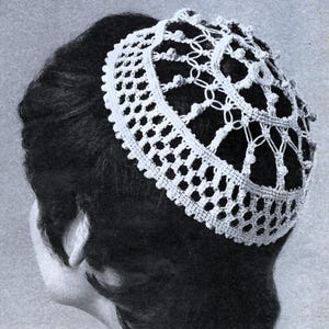 May include: A white, crocheted head covering, possibly a doily or a hair accessory, is worn on the head. The intricate openwork design features a repeating pattern of small squares and circles, with a scalloped edge. The head covering is set against dark hair.