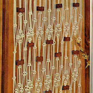 May include: A macrame wall hanging featuring a repeating pattern of knotted white cords and rectangular wooden beads. The design is framed by a dark wooden border, creating a textured, bohemian aesthetic.